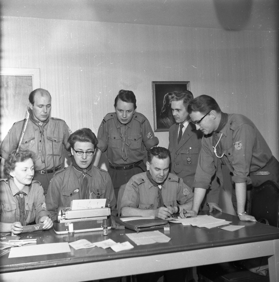 Sex män och en kvinna befinner sig vid ett skribord. Tre personer sitter, varav en sitter framför en skrivmaskin. Fyra personer står lutade över de sittades.