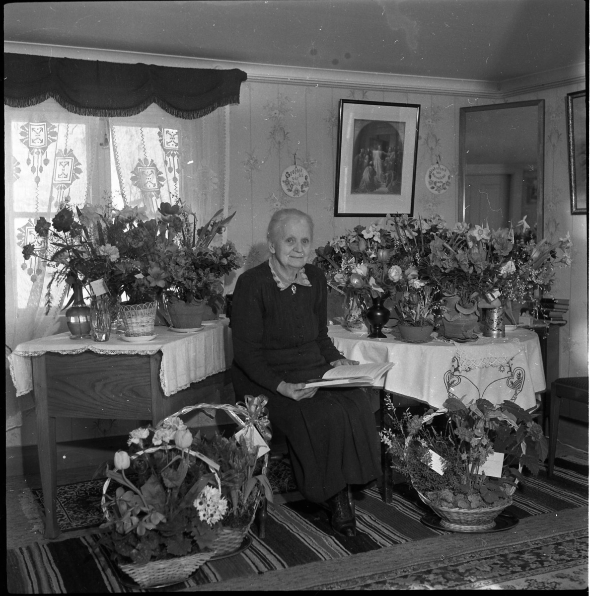 En kvinna sitter omgiven av blombuketter och blomsterarrangemang. Hon håller i en bok. Hon bär en svart klänning med spetskrage.
