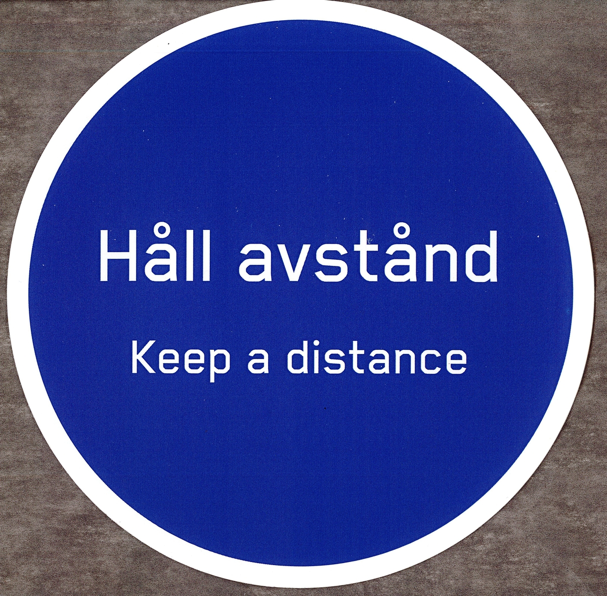 Blå rund klisterdekal med texten ”Håll avstånd Keep a distance” i vitt. Vit kant runt om. Vit baksida.