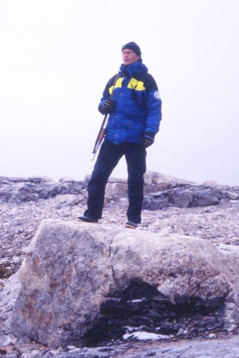 En man i Polarforskningssekretariatets kläder med gevär över axeln står på en större sten. Han är björnvakt i samband med utgrävning av Andréelägret på Vitön.
