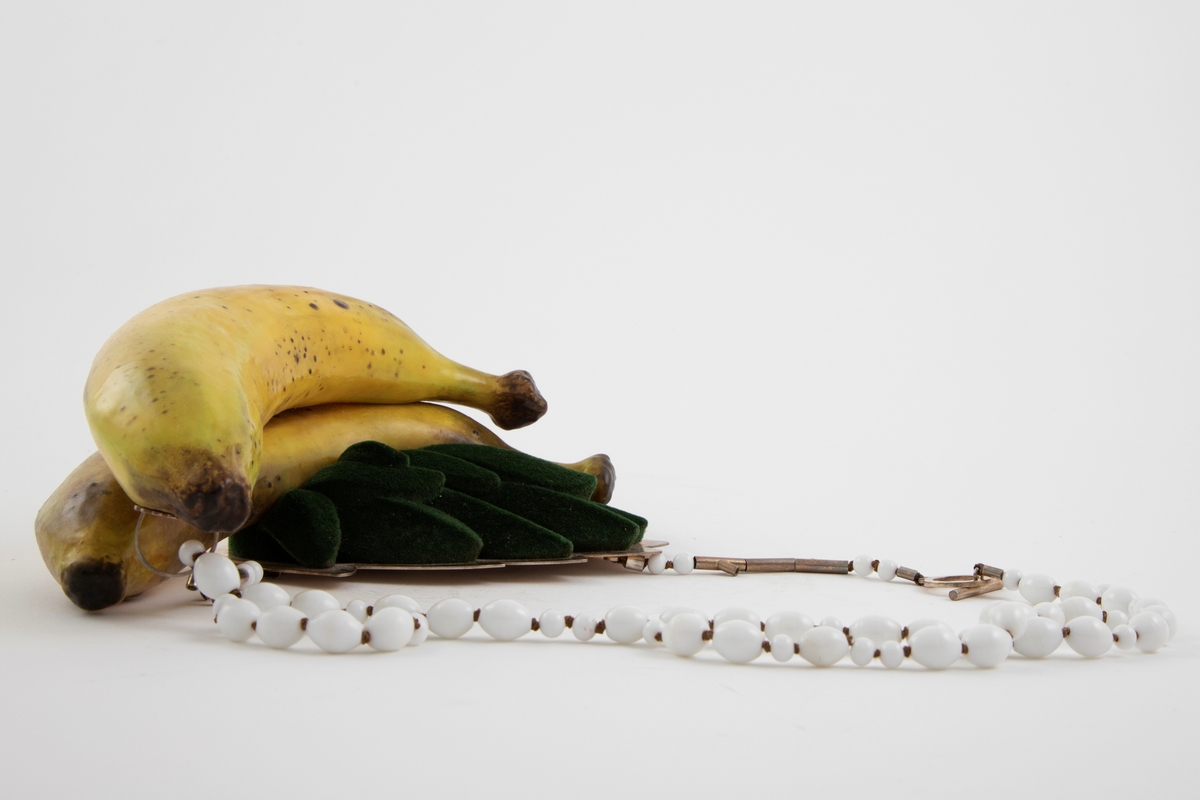 Skulpturell halssmykke med to naturtro bananer i gummi og blader av pålimt grønn flokk (minner om velur), festet på en sølvplate. Kjede med hvite glassperler i ulik størrelse og enkel lås i sølv.