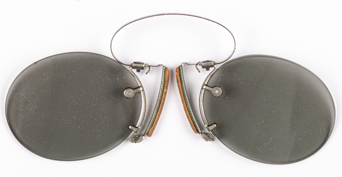 Pincené, ovala gråfärgade ögonglas utan fattning. "Näsklämman" av vitmetall, försedd med gummilist. Ögonglasens L=4,2 cm, B=3,3 cm.