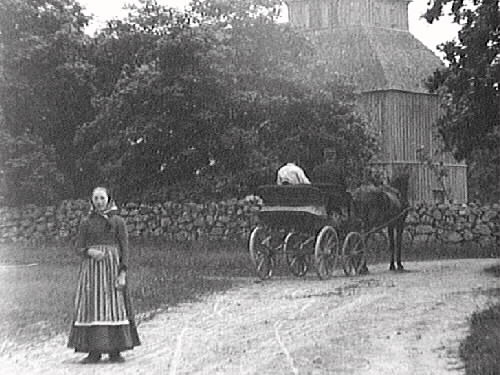 En hästskjuts är på väg mot Okome gamla kyrka och i förgrunden står en kvinna med randigt förkläde vänd mot fotografen. Okome nya kyrka uppfördes 1891.