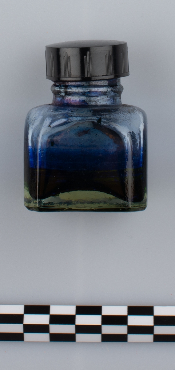 Glassflaske med tørkede rester av formentlig blått blekk. Sort skrulokk i kunstmateriale. Etikett på en side.