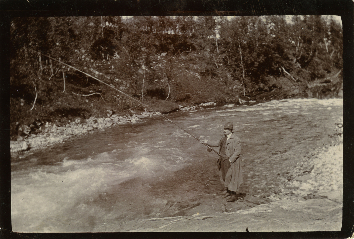 En mann med med mustasje, iført lang frakk og hatt står ved elva og fisker. Venstre hånda har han puttet avslappet i bukselomma mens han holder fiskestanga med høyre hånd.