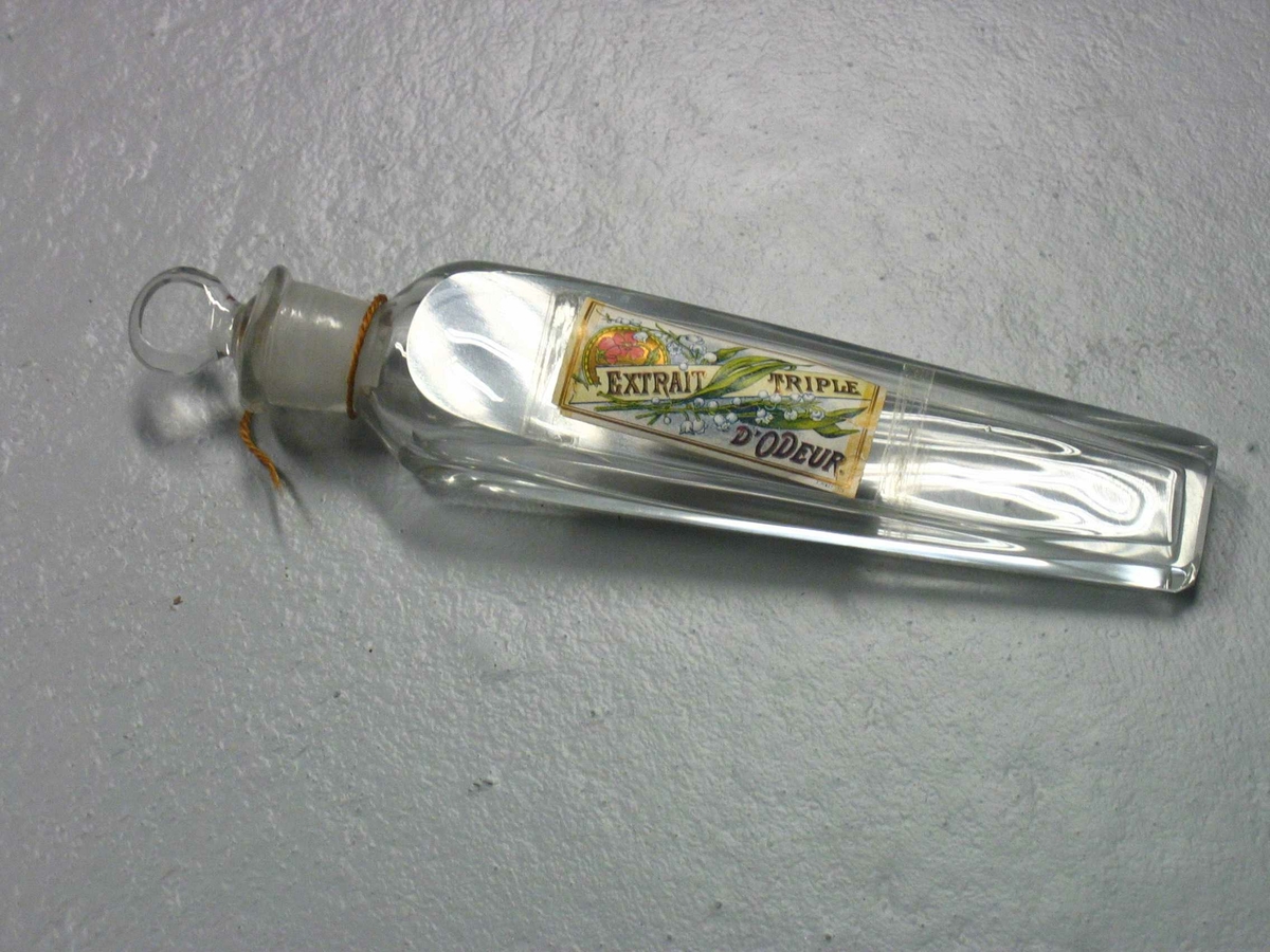 Avlang parmymeflaske av glass med propp. Pålimt papiretikett med tegning av liljekonvalller og andre blomster og teksten EXTRAIT TRIPLE D'ODEOUR