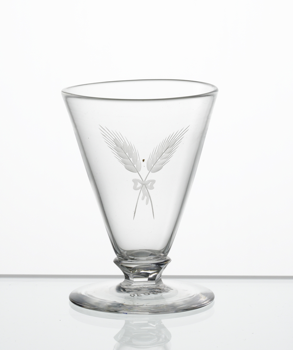 Design: Edward Hald.
Brännvinsglas, konisk kupa med fasettslipad knapp mellan kupa och fot. Graverat motiv i form av korslaga sädesax sammanbundna med en rosett, på kupan.