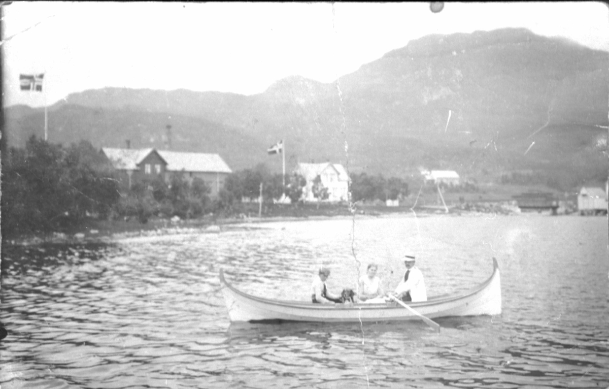 Tre personer med hund i en nordlandsbåt. I bakgrunnen ser vi Sørvik Meieri.
