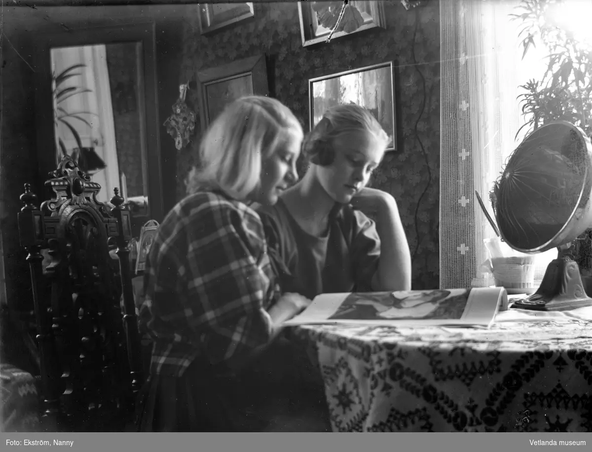 Två flickor vid ett bord. Flickan närmst kameran är Anna Karin Zackrisson. Anna Karin var dotter till fotografen Nannys syster Amy. Bilden är troligtvis tagen i fotografens bostad på Kyrkogatan.