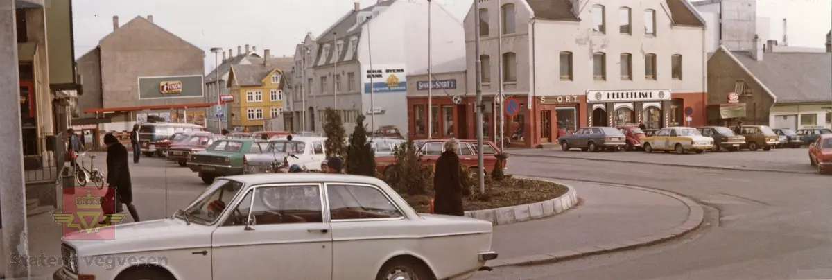 Haugesund i Rogaland april 1977. Mange biler står parkert. Fremst i bilde står en hvit Volvo 242. I bakgrunnen en hvit Peugeot 404, og en grønn Ford Taunus. På andre siden av gaten står blant annet en hvit og gul Ford Cortina og en rød  Volkswagen Boble.
