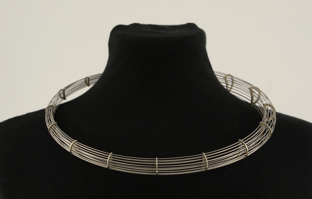 Halssmykke laget av 16 tynne metalltråder som er tredd gjennom 19 halvmåneformede metallstykker. Metallstykkene minsker i størrelse, slik at smykket smalner i den ene enden. Smykket danner en ring ved at den smale enden er tredd inn i den brede.