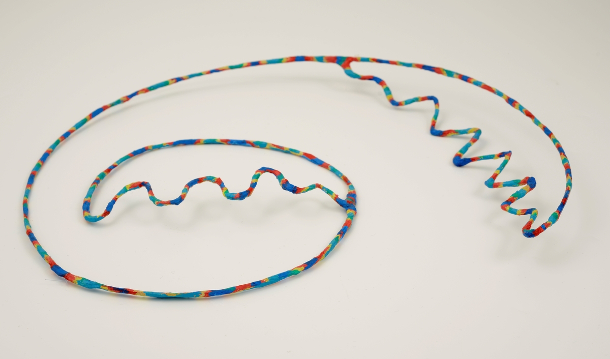 Halssmykke laget av metalltråd som er dekket med papir malt med akrylfarge i forskjellige farger; rød, blå, gul og grønn. Smykket er formet som en åpen spiral som ender i siksakk-former ytterst og innerst.