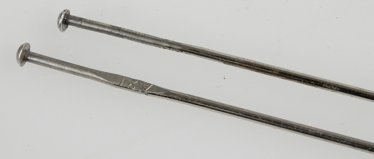Ett par stickor av metall, under knoppen en utbuktning med text: AXT på ena sidan och siffran 2 på andra sidan.