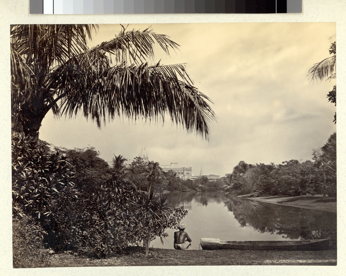 Bilden visar Calcutta äldsta parkanläggning Edens Garden. I förgrunden syns en man bredvid en båt som tittar ut över en vattenyta. I bakgrunden ser man byggnaden av Calcuttas högsta domstol.