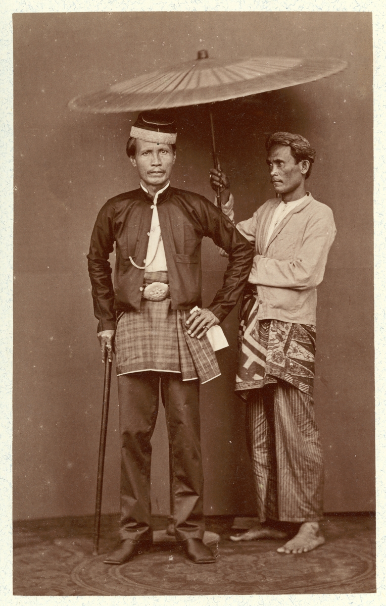 Bilden visar två malajer som verka vara herre och betjänt. Mannen till vänster bär påkostade kläder, huvudbonad, fina skor och ett käpp. Bredvid honom står en man i enkla kläder som är barfotat och håller en paraply över honom.