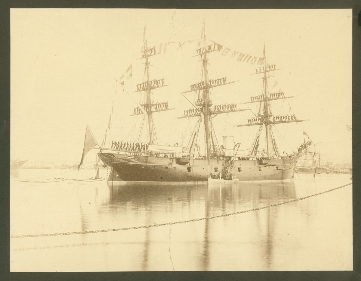 Bilden visar en råmanning på korvetten Freja. Fartyget ligger till ankars i hamn, alla segel är revade. Hela manskapet står på däcket och ovanpå rårna och hälsar med sina mössor. Dessutom bär fartyget storflaggning.
