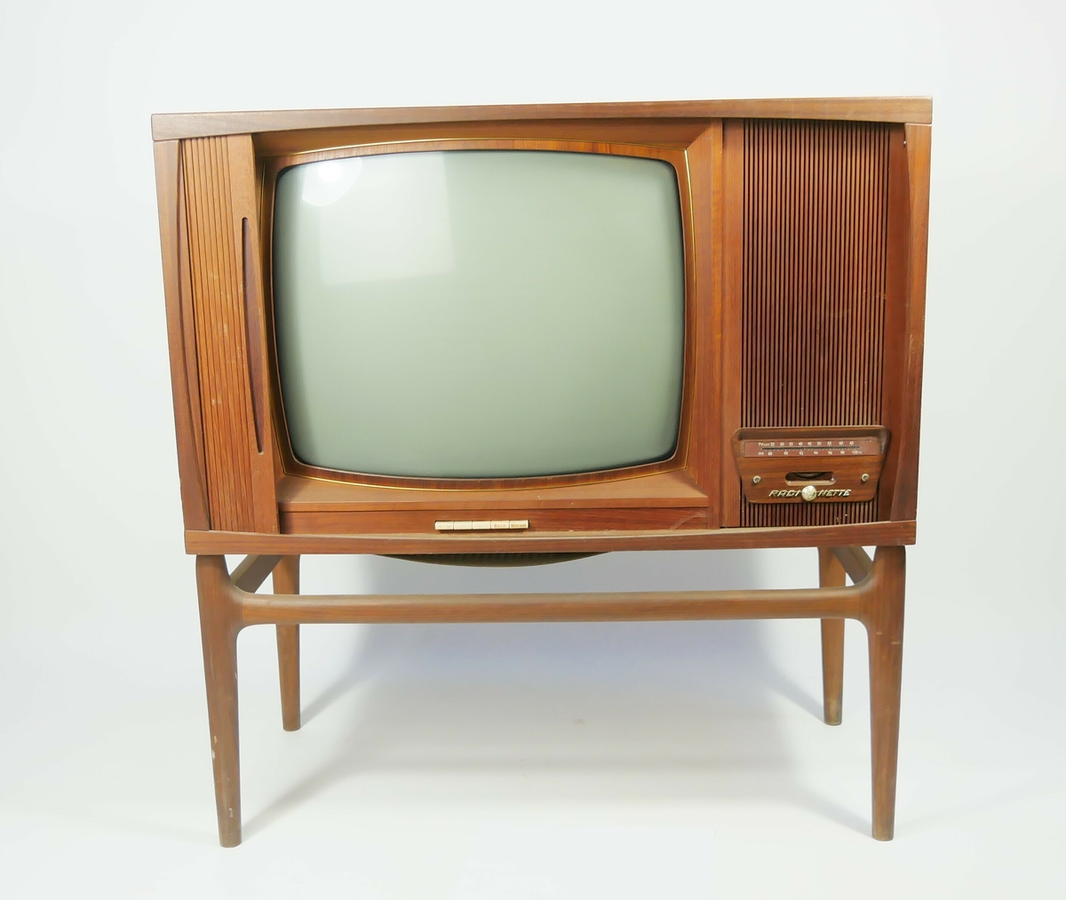 Rektangulært fjernsynsapparat som står på fire bein. Foran på den en siden er det en treskjerm foran tvskjermen som kan skyves til siden. 