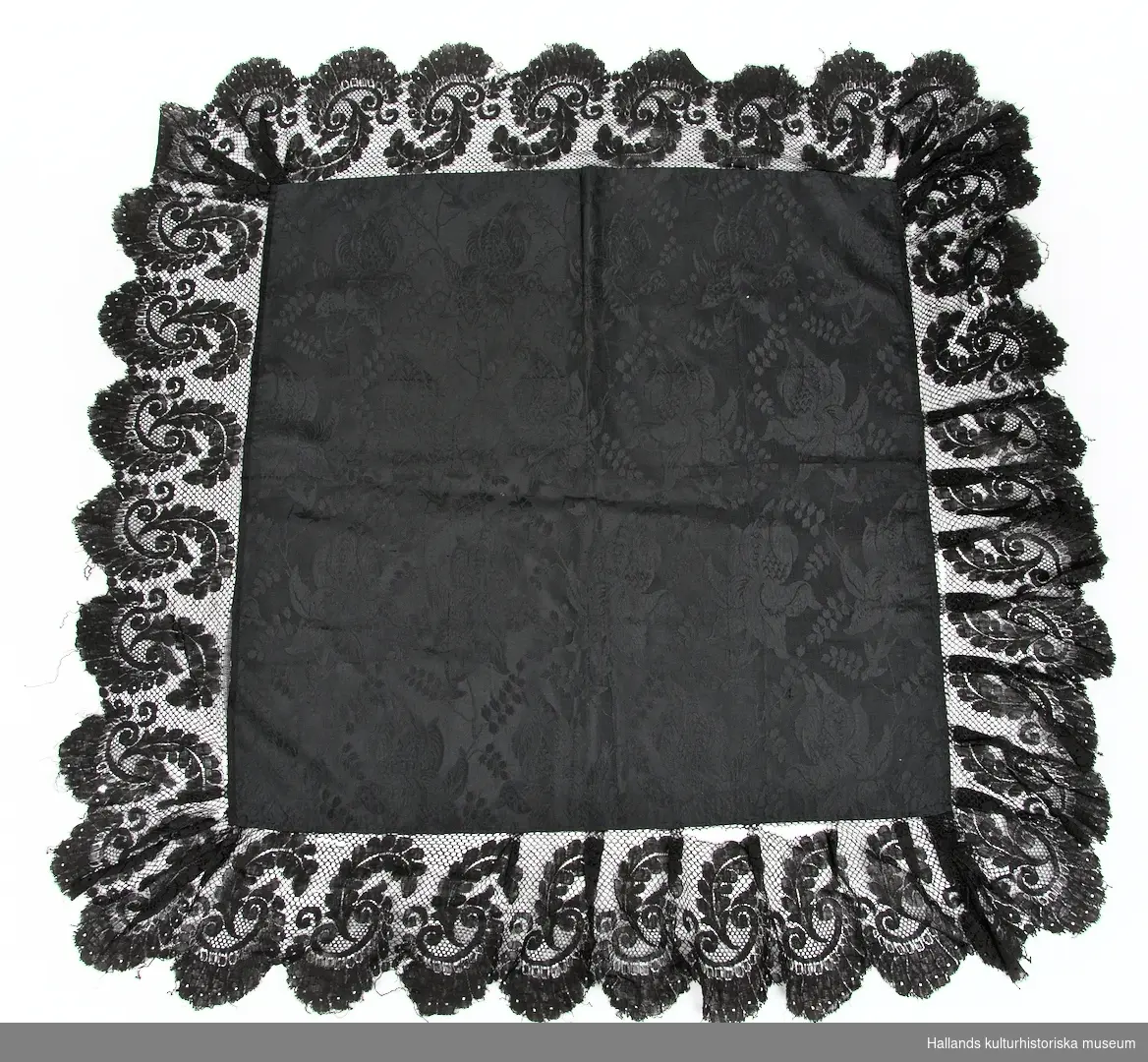 Sjal i svart sidentyg med invävda storblommiga mönster, dekorerad med en cirka 15 cm bred spetskant i ett gemoetrisk blommönster.