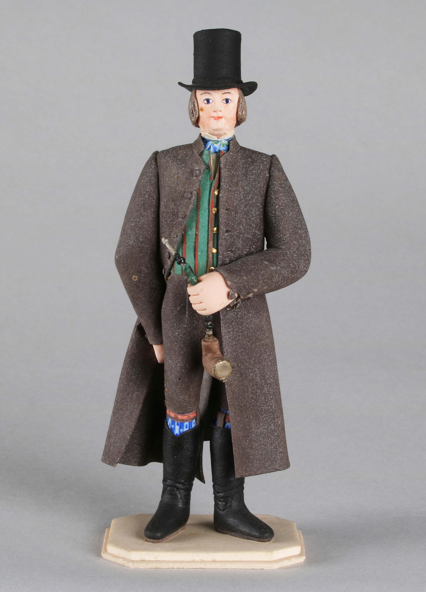 Dragantarbete. Mansfigur.
Allmogedräkt. Stående mansfigur, iförd hög svart cylinderhatt. Trekvartslång, gråsvart rock med långa ärmar. Grön- och brunrandig väst och mörka knäbyxor. Svarta stövlar som räcka till knäna. Strumpor, mönstrade, blå och röda samt strumpeband. Blå knuten halsduk. Mannen bär en stor pipa i vänster hand. Står på åttkantig ljus platta.

Tillverkad av Hilda Boström (1822-1903), sockerbagare, Borås.

Litt: Se Årsbok 1963, sidan 45.

Funktion: Prydnadsfigur