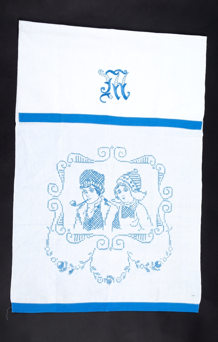 Pyntehåndkle, Hvit bunn, blå kanter. Brodert med en gutt og ei jente med korsting. Monogram M.

Gitt av Erna Madsen. Produsert av Margit Madsen.
Disse drev manufaktur i Drøbak.