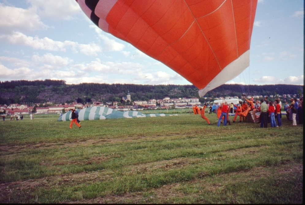 En orange-blå-vit ballong står klar att lyfta. Ballongen lutar kraftigt åt vänster. Personer i orangea overaller håller i ballongen.