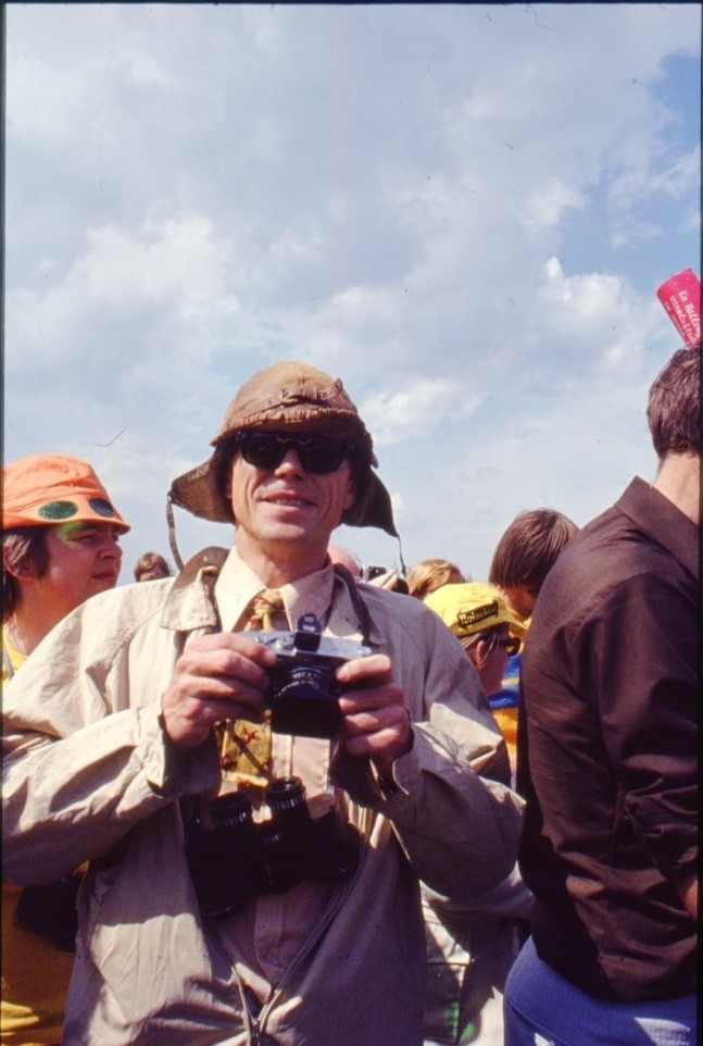 En man i mössa med öronlappar och kamera i händerna står vänd mot fotografen, omgiven av andra människor.