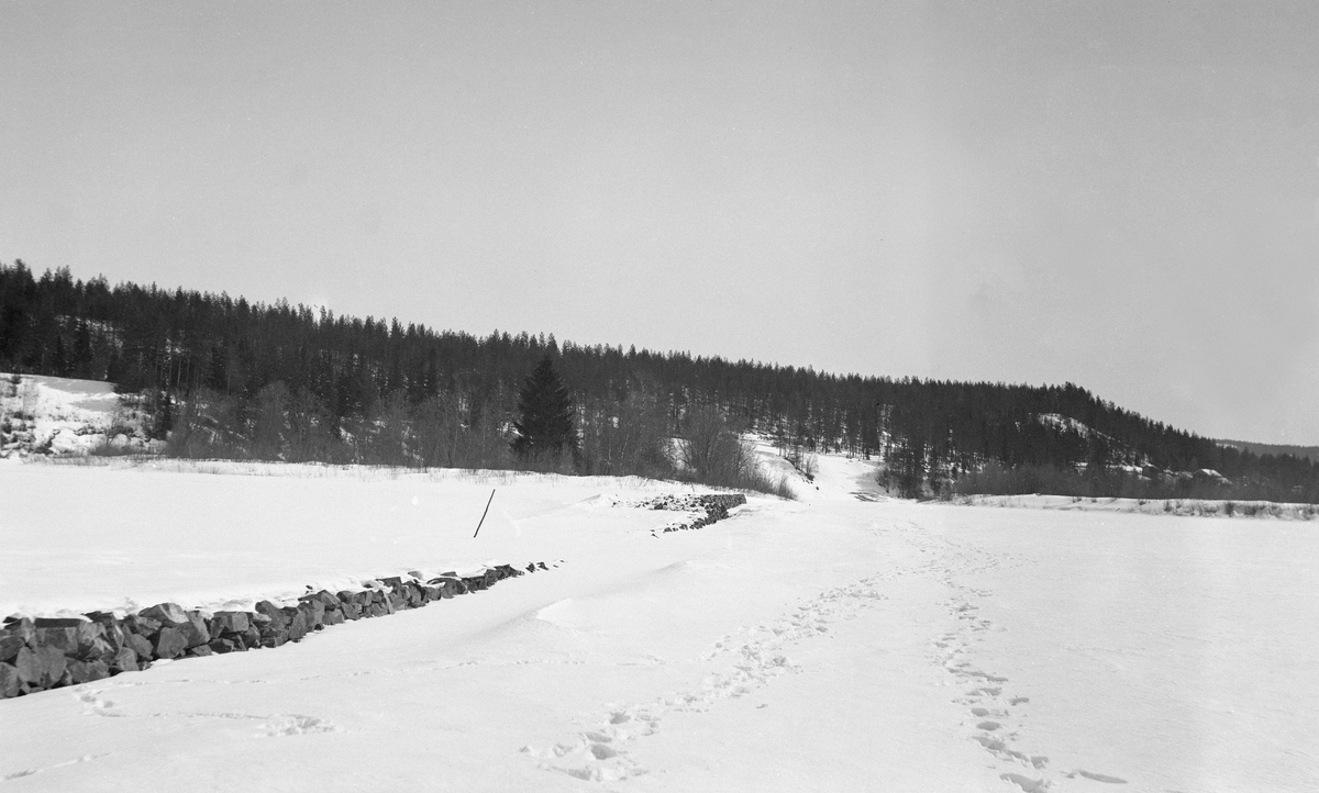 Vassdragsvesenets forbygninger mot isgang ved Alme i Åmot, fotografert vinteren 1933.  Fotografiet er tatt fra ei flat, snødekt slette, antakelig isen på Glomma.  Til venstre i bildet ses et slags steingjerde, som skal være opplagt som en forbygning mot blant annet isgang i elva.  Deler av forbygningen er dekt av snø, noe som i underteksten under albumkopien av fotografiet tas som et tegn på at muren er for lav.  I bakgrunnen en bakkekam med barskog. 