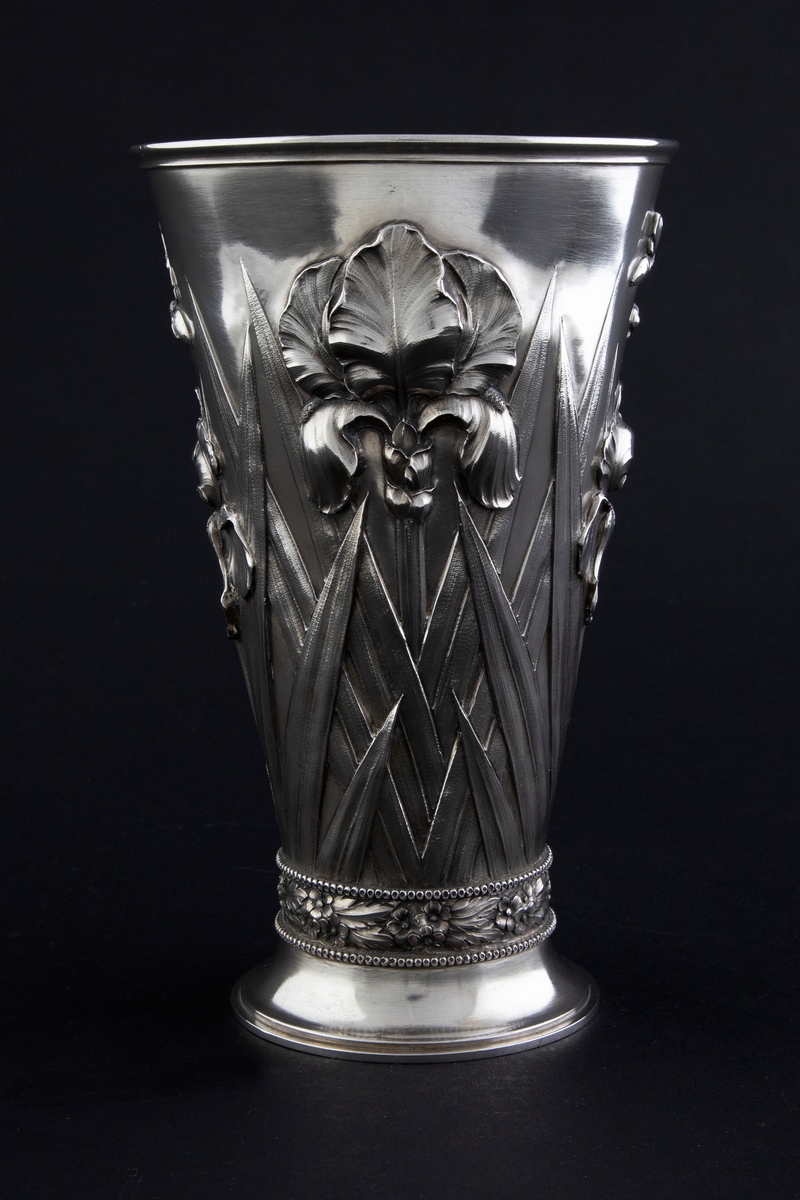 Begerformet vase i sølv. Konisk korpus som er dekorert med drevede irisblomster, omgitt av stiliserte planteformer. Overgangen mellom korpus og den hevede foten er markert med en frise av blomsterornamenter i relieff. Innsiden er forgylt.