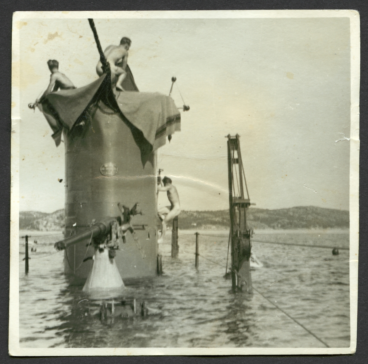 Bilden visar ubåten Najad vars däck ligger under vatten, bara tornet och överbyggnader sticker ut. På tornet syns tre nakna ubåts soldater, den ena håller på att klättra upp på sida av tornet. Runt omkring i vattnet syns fler besättnings män som badar.