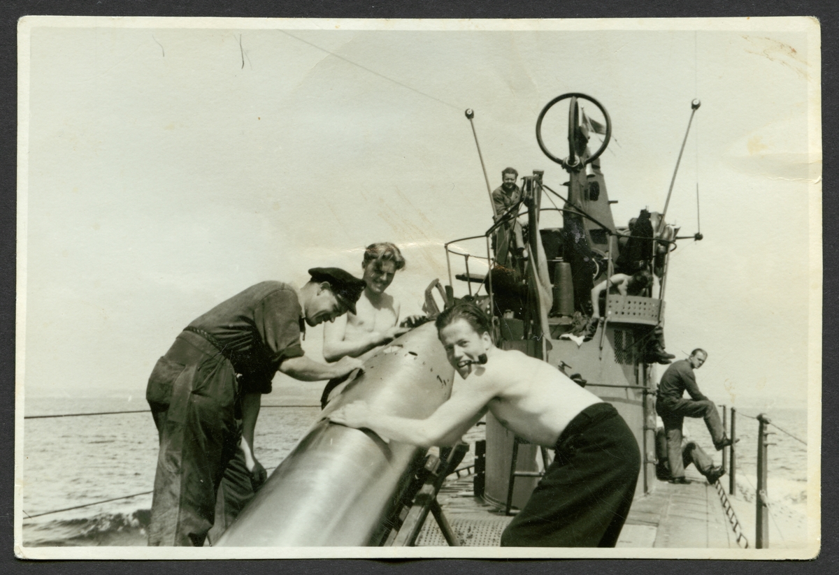 Bilden visar tre sjömän som arbetar med en torped på däck av ubåten Najad. En man bär arbetskläder de två andra arbetar med bar överkropp och mannen in förgrund röker en pipa.