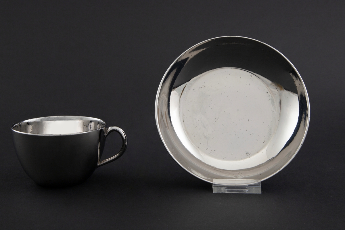 Kopp med skål i keramikk, glasert med sølvfarget luster. Tilnærmet sylinderformet korpus med påsatt hank. Lav sirkulær skål.
