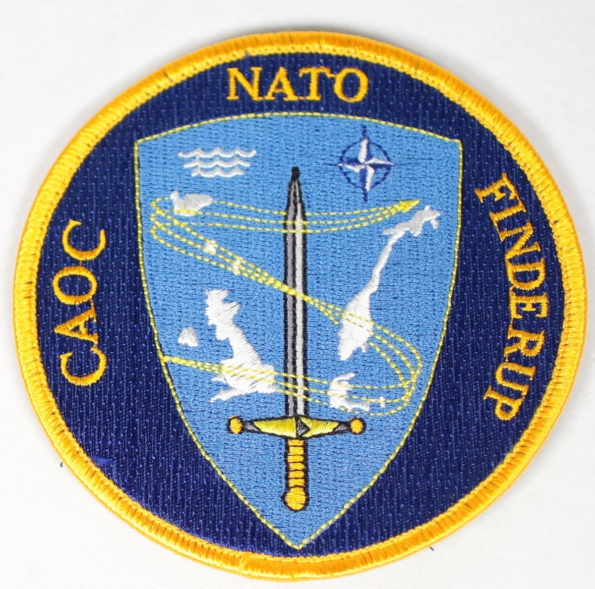 The insignia for Combined Air Operation Centre Finderup has, at its centre, the sword of Component Command Air Ramstein. Component Command Air
Ramstein is the Headquarters responsible for all NATO air activities within the Northern Region. To the left and right of this protective sword are the nations: Iceland, United Kingdom, Denmark and Norway, within whose airspace all NATO air activities is the responsibility of CAOC Finderup – most important the air policing by NATO Quick Reaction Alert assets as indicated by the aircraft symbol. Three aircraft tracks merge into one to symbolize the amalgamation of former CAOC Finderup, CAOC Reitan (Norway) and CAOC High Wycombe (UK) into CAOC Finderup.