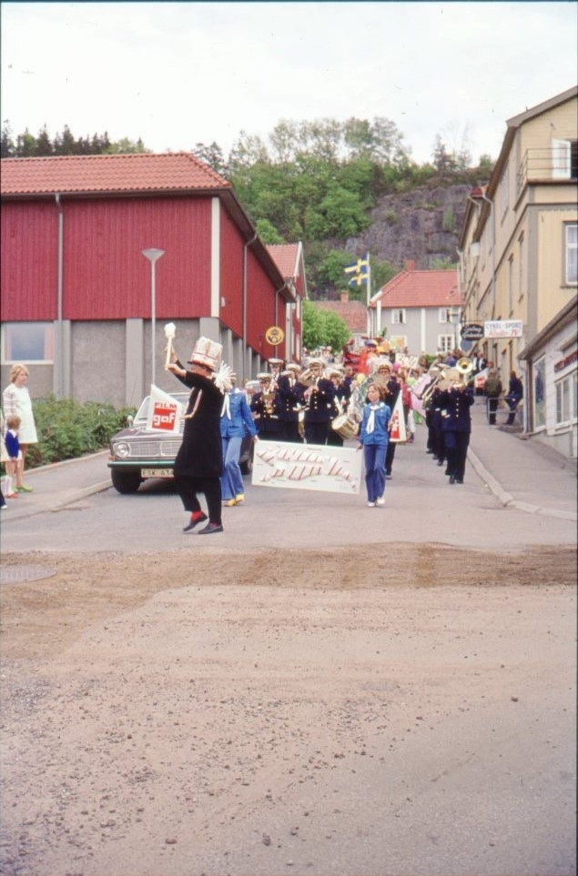 En musikkår kommer gående nedför Ribbagårdsgränd för att förmodligen snart svänga norrut på Sjögatan. I täten går en man i svart och med spexig hög hatt och röda strumpor som tamburmajor.