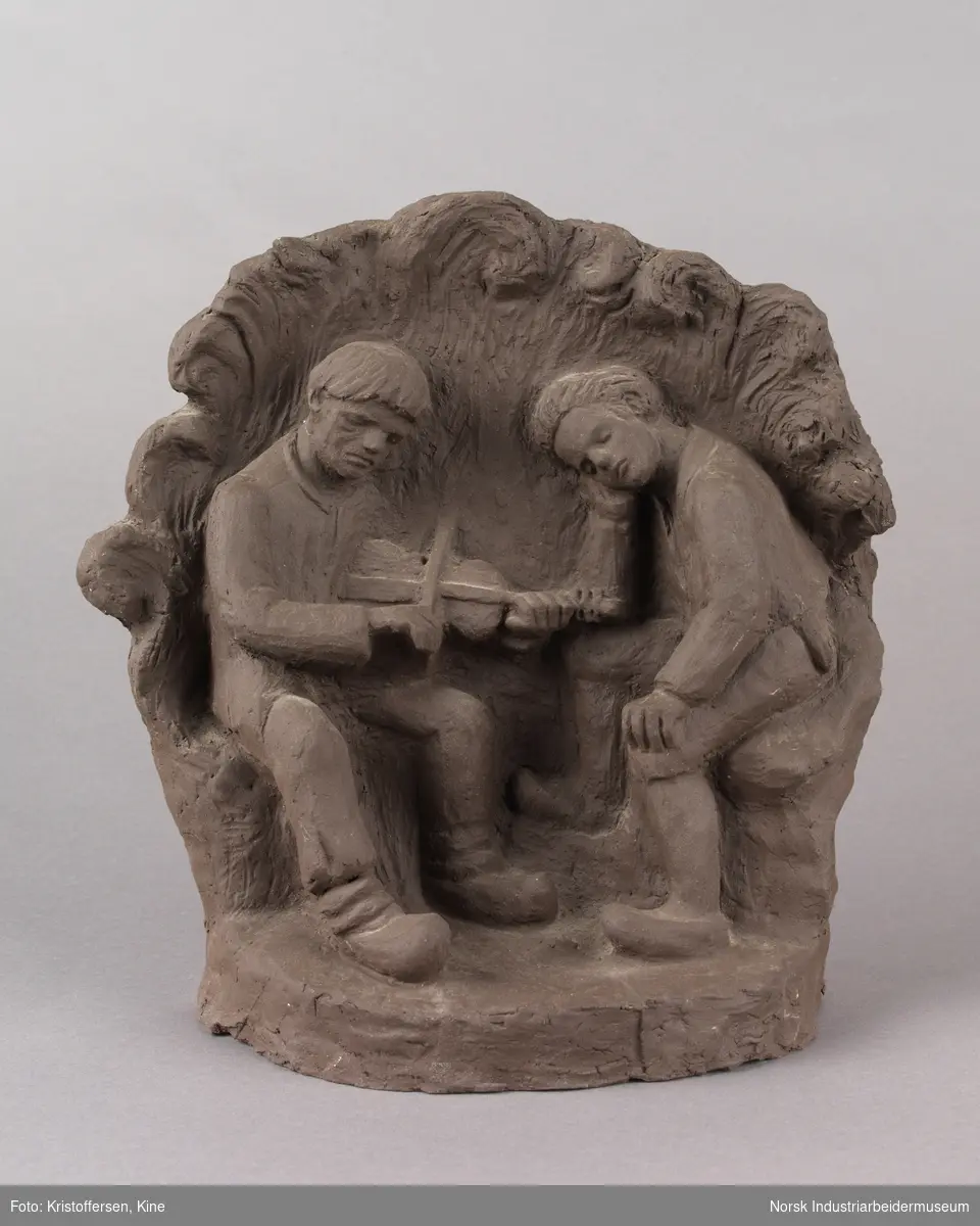 Skulptur laget av leire. En spelemann spiller på en fele, mens en tilhører sitter ved siden av. Tilhøreren har en hand på låret og luter hodet mot den andre handen. Skulpturen består av to sittende figurer, en grunn og en bakgrunn med bølgende kanter.