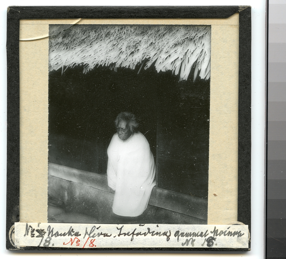 Bilden visar en äldre kvinna på Nuku Hiva klädd i en traditionell dräkt som sitter i ingången till ett trähus. Enlig Etnografiska museets bildinformation är bilden tagen på Fakarava.