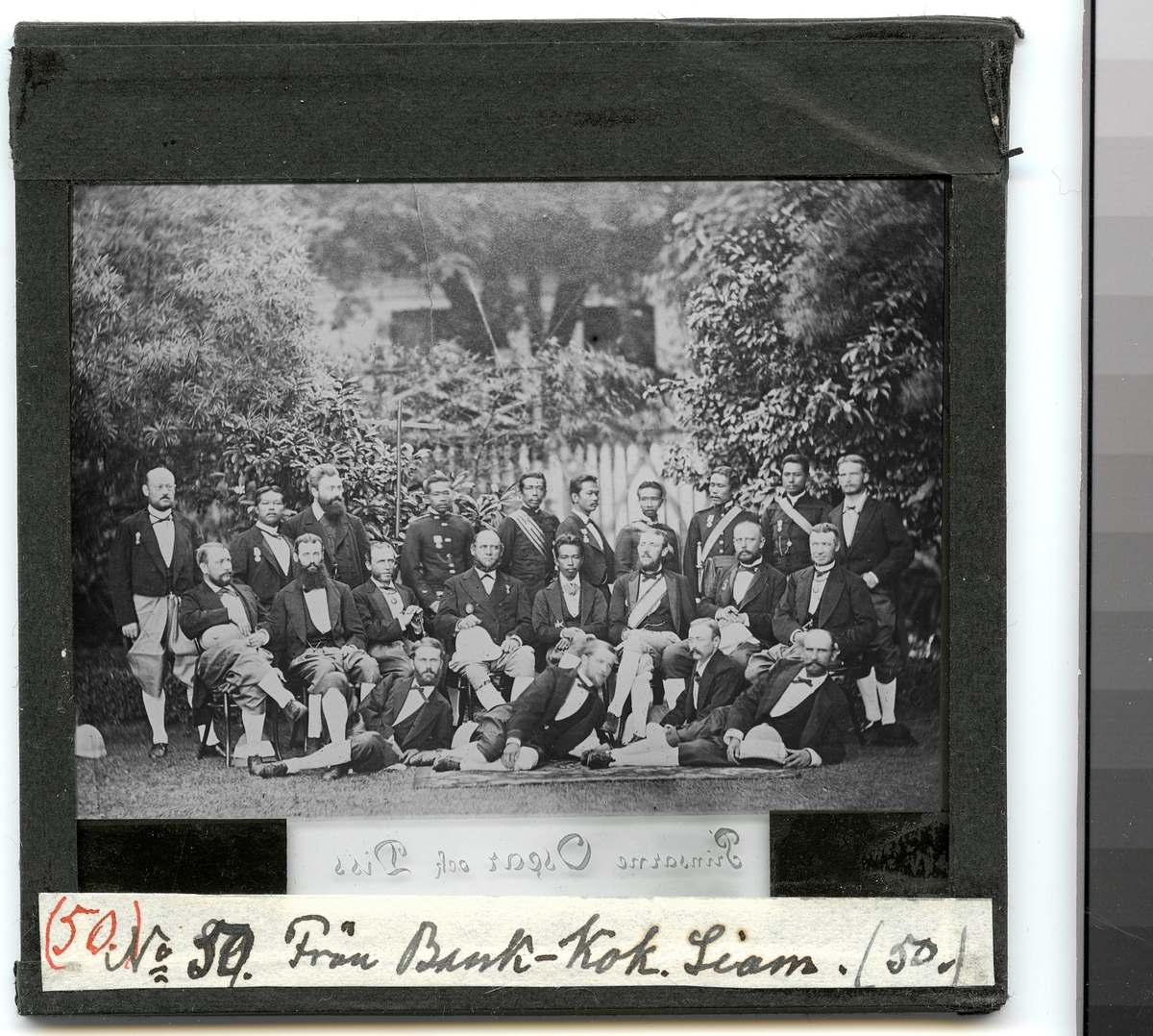 Denna grupporträtt visar officerare från fregatten Vanadis och prins Oscar Bernadotte tillsammans med siamesiska hovman som har samlats för fotografering i en trädgårdsmiljö. Alla bär siamesiska dräkter.