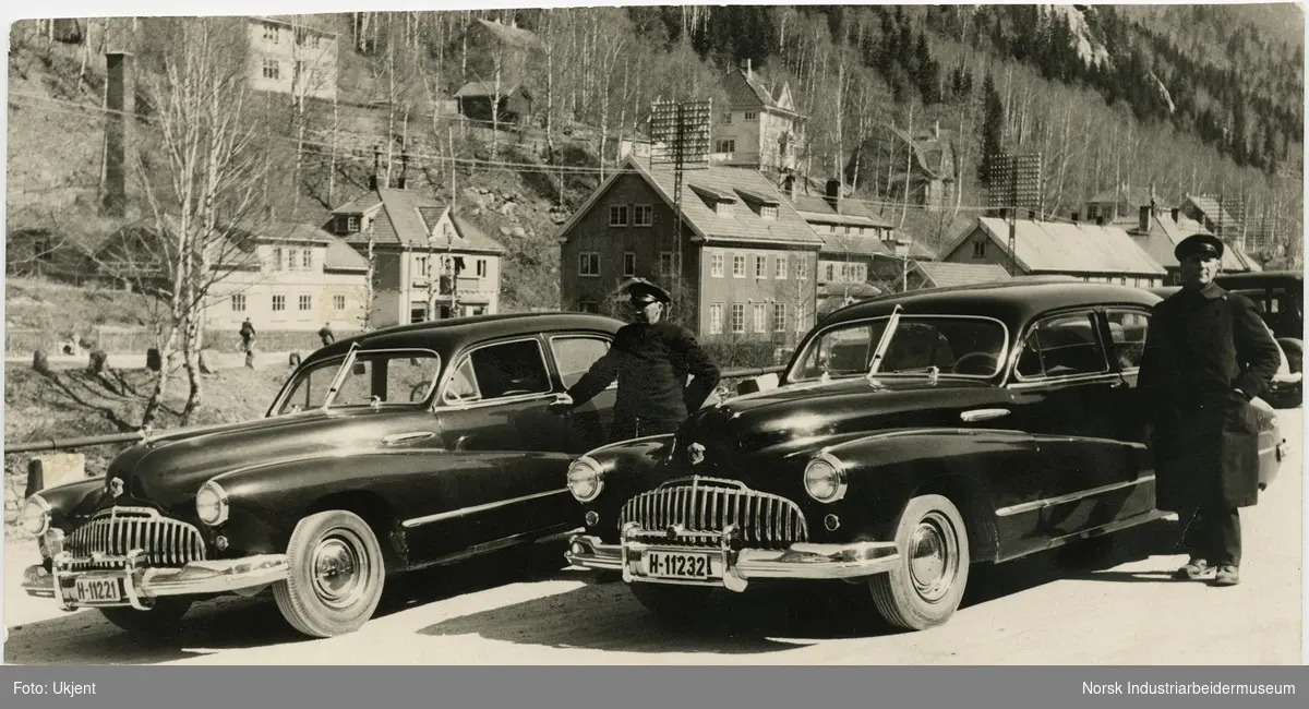 Oskar Andresen og Ole Brenden i uniform stående ved siden av hver sin bil av merket Buick. Bilene tilhører Rjukan Byanlegg og mennene var ansatt som sjåfører i Norsk Hydro. 
Rjukan Varelager, slakter og bakeri i bakgrunnen. Boliger i Villaveien.