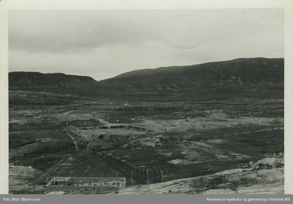 Ødeleggelser i Fuglenesdalen i Hammerfest. I forgrunn ser man et inngjerdet område og ruiner av hus. Små veier krysser området. I bakgrunn ligger Vardfjellet til venstre og Storfjellet til høyre. Bildet er mest sannsylig tatt fra Fuglenesfjellet. Det er skrevet "Fuglenesdalen" på baksiden av bildet.