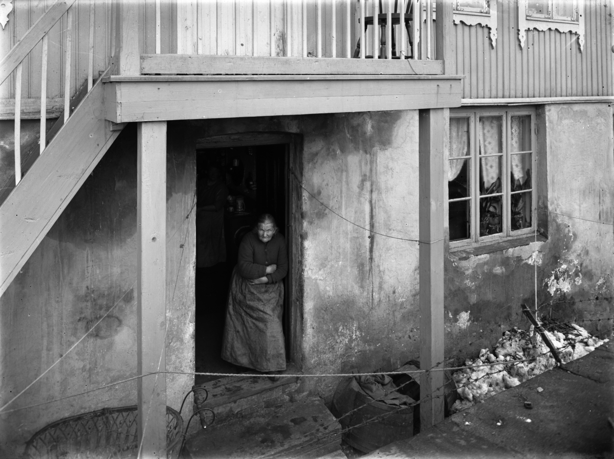 Gammel kone betrakter fotografen fra døråpningen i slitt hus. Piggtråd foran døra.