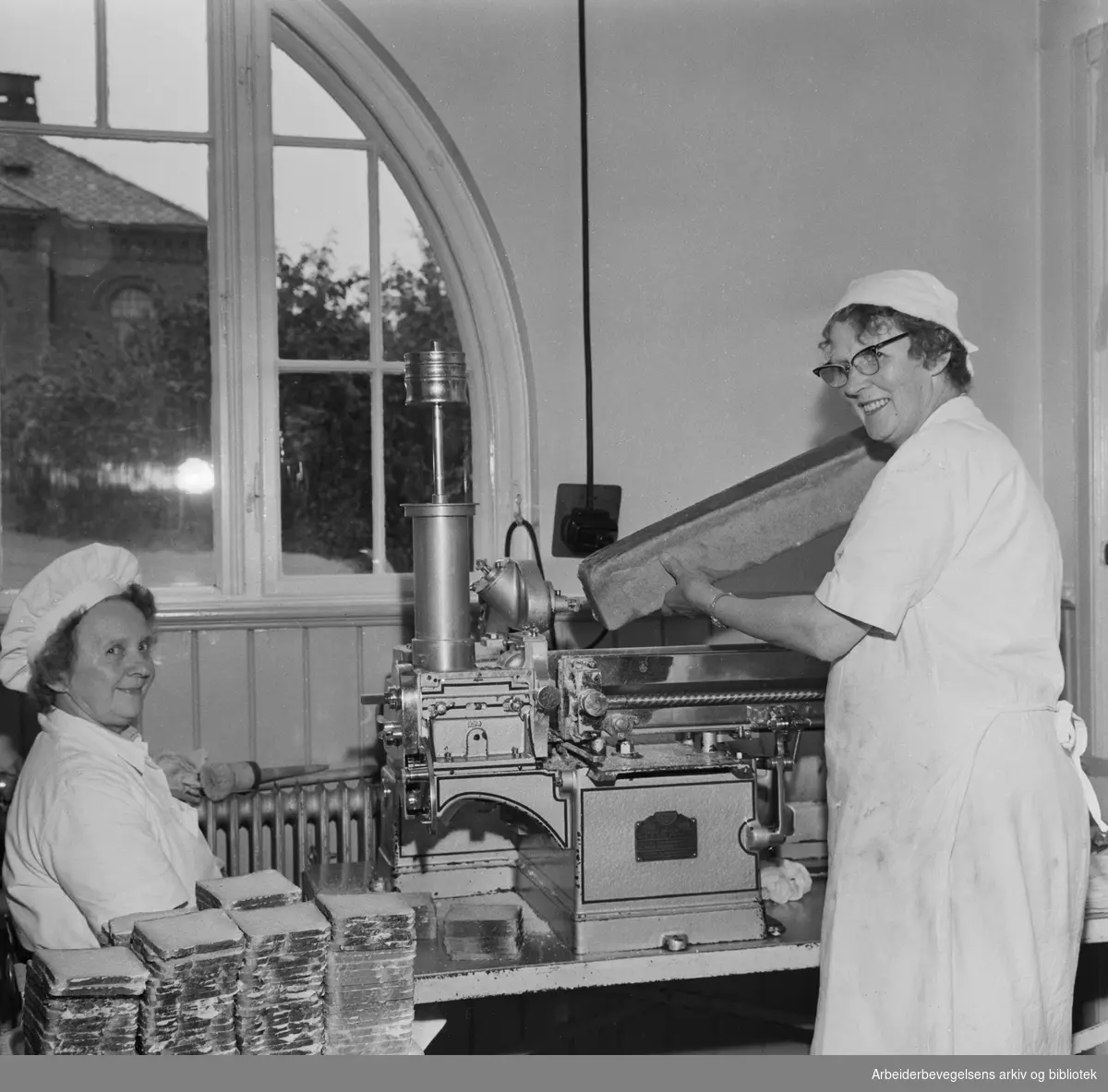 Oslofrokosten. Astrid Krogh og Ragnhild Engh ved den automatiske smørbrødmaskinen på Oslo kommunes kjøkken i Lakkegata. September 1960