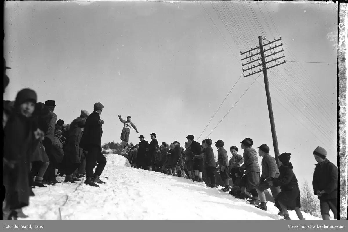 Jernbanens idrettslag arrangerer hoppkonkurranse. Folkemengde samlet i bakken for å se skihopp. Utøver nr 20 i lufta.