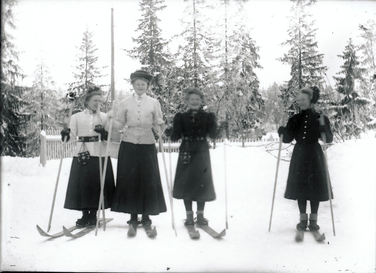 Fire unge kvinner på ski. To og to i nesten like drakter. Kvinnene til venstre i lyse strikkejakker med belte i livet og ankellange, mørke skjørt. Kvinnene til høyre i mørke jakker med belte, mørke skjørt til midt på leggen og raggsokker brettet over støvler.