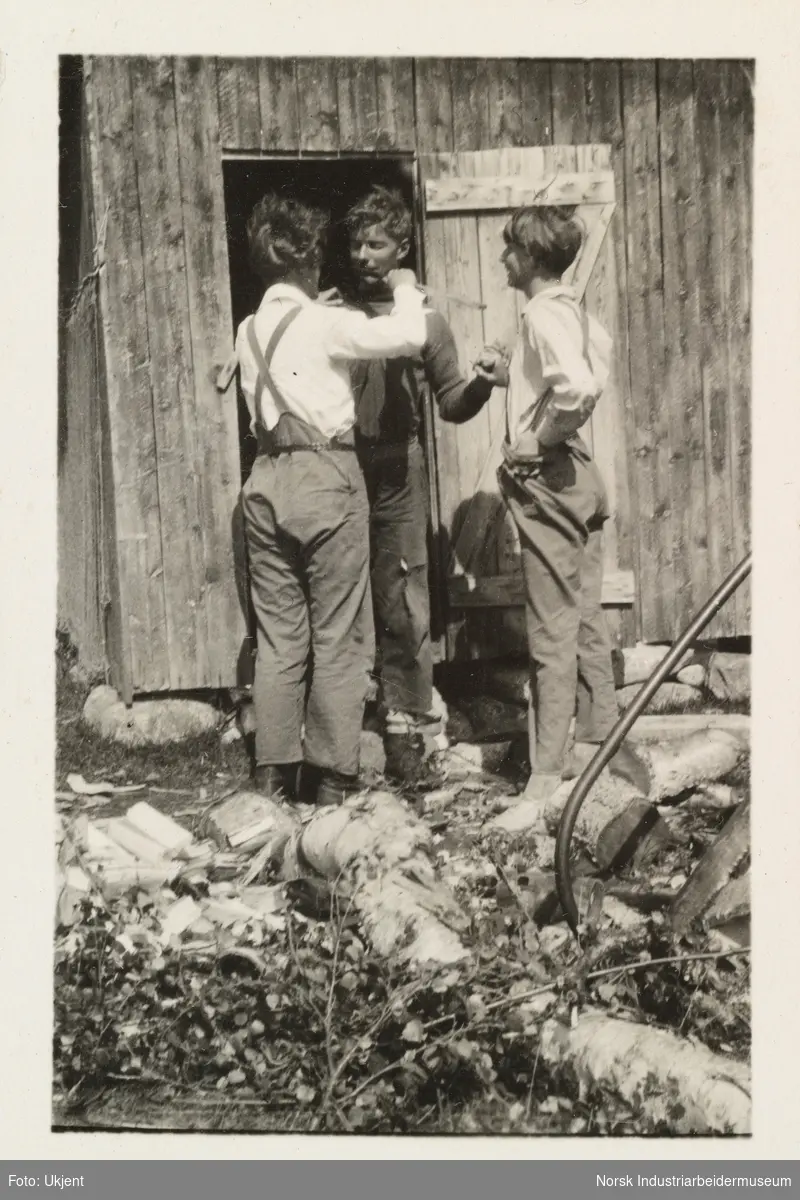 Kvinne holder noe foran munn på en mann som står i døråpningen på et uthus på Møsstrond. Mannen holder den andre personen i hånden. I forkant tømmer, ved og sag