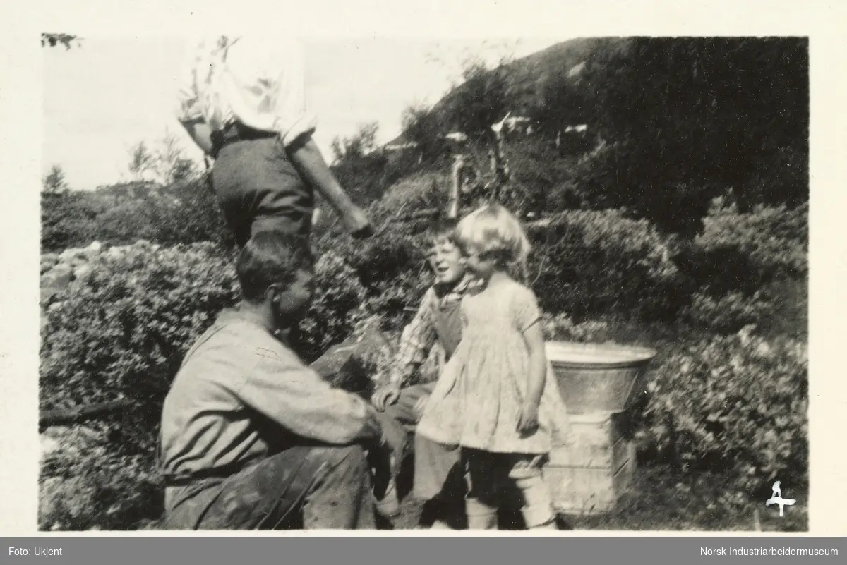 Mann sitter på huk foran to små barn utendørs på Briskeroe, Møsstrond. Bak sees en vaskebalje på en trekasse