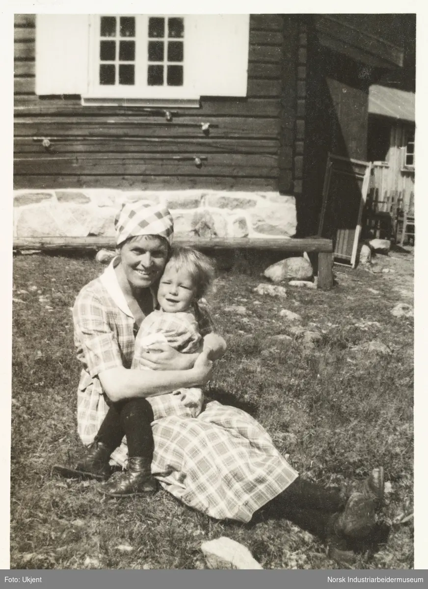 Anna Poulsson sitter på gresset med Sven Hartvig Poulsson på fanget foran hytta på Briskeroe, Møsvatn