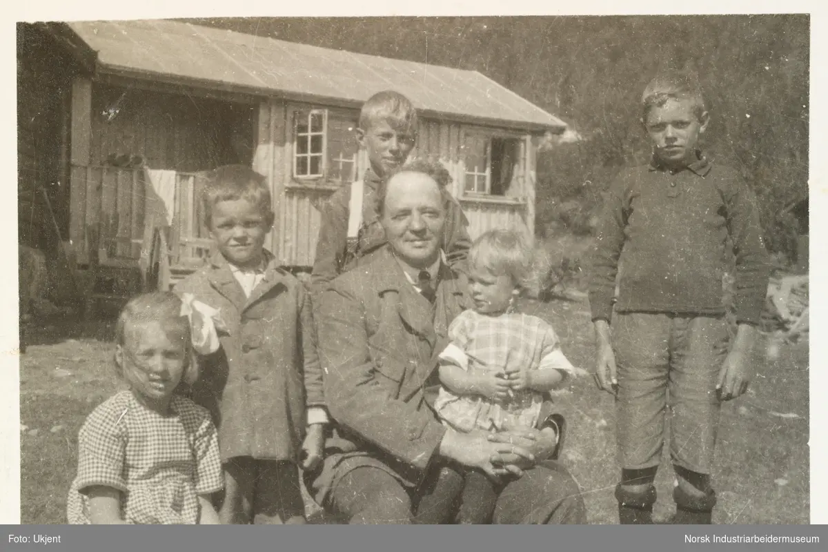Briskeroe, Møsstrond. Magnus Poulsson sitter foran hytta med fem barn. Ellen Poulsson sitter på fanget til faren Magnus. Fra venstre: Berit, Anton, Magnus, Esben