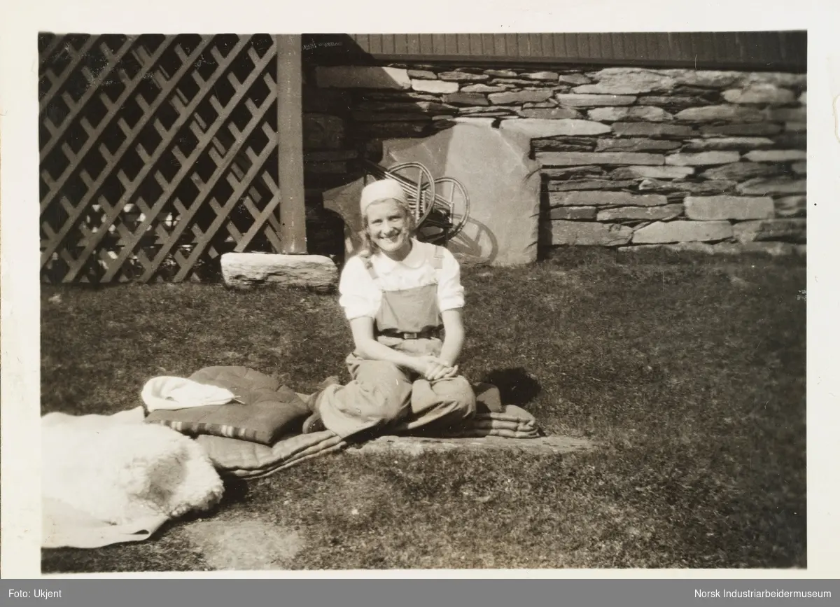 Sommeren 1931, Gullia og Fagerli. Kvinne i snekkerbukse og skaut sitter utendørs på puter og pledd på gress ved bolighuset på Gullia, Lysaker