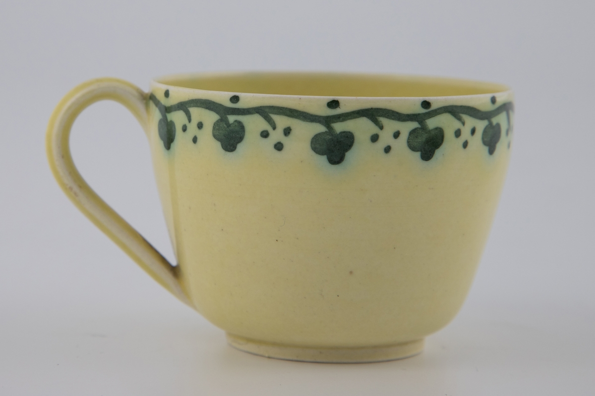 Kaffekopp med tilhørende skål i gulfarget glasur, dekorert med emaljemaling. Dekoren er mørkegrønn, og er utformet som et stilisert planteornament som slynger seg langs fatets og koppens kant.