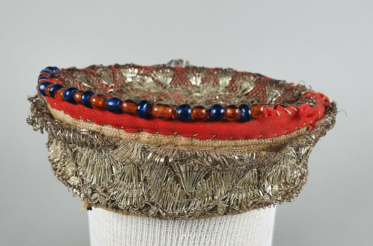 Valk for brudepike, trukket med lin og rød ull. Rund kanten og på toppen er det påsydd en sølvblonde av metall og rund deler av kanten er det påsydd perler i blått og brunt.