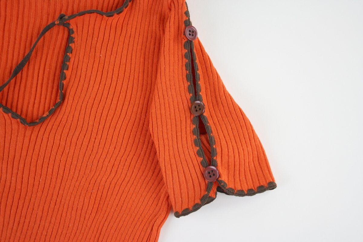 Kortärmad orange tröja.
Tröjan har en snörning i urringningen och en resår som bildar en volang vid midjan. Ärmarna har en slits som är ihopfästad med tre bruna knappar. Halslinning, snören samt kanterna är bruna. 
Tillverkad av Fix i storlek 150.
